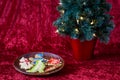 Fresh baked sugar cookies decorated by children for Christmas treats, on a wooden plate, with lighted tree, on a red background Royalty Free Stock Photo