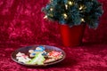 Fresh baked sugar cookies decorated by children for Christmas treats, on a wooden plate, with lighted tree, on a red background Royalty Free Stock Photo