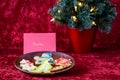 Fresh baked sugar cookies decorated by children for Christmas treats, on a wooden plate, with lighted tree, on a red background Royalty Free Stock Photo