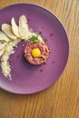 Fresh beef steak tartare with parmesan cheese, capers, croutons and yolk quail on a purple plate on a wooden background