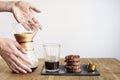 Fresh coffee in Chemex, sweet brownie cookie on wooden table. Beautiful woman`s hands pouring from glass bowl. White wall behind