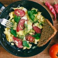 Fresh cooked scrambled eggs in pan with sausage and herbs. Bread, tomato, chilli pepper on wooden board, top view. Royalty Free Stock Photo