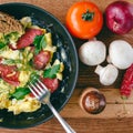 Fresh cooked scrambled eggs in pan with sausage and herbs. Tomato, onion, mushrooms on wooden board, top view Royalty Free Stock Photo