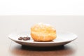 Fresh doughnut with icing sugar and coffee beans on white ceramic plate on bright light brown wooden table