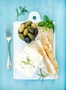 Fresh feta cheese with olives, basil, rosemary and bread slices on white ceramic serving board over bright turquoise Royalty Free Stock Photo