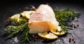 Fresh fish, raw cod fillet with addition of herbs and lemon slices on black stone background