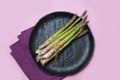 Fresh green asparagus on black ceramic dish, purple linen napkin on pink background.