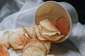 Fresh homemade deep fried crispy potato chips in white box on a wooden tray, top view. Salty crisps scattered on a table