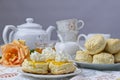 Fresh scones topped with apricot jam and whipped cream with tea pot and cups in background
