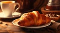 Fresh hot croissant on a white saucer and a cup of coffee next to on wooden surface, French classic breakfast, close-up.