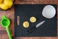Fresh lemon cut in half on a black cutting board, paring knife, wood table, basket of lemons, citrus squeezer, bowl