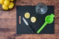 Fresh lemon halves on a black cutting board, paring knife, wood table, basket of lemons, citrus squeezer, drinking glass