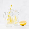 Fresh lemonades with lemon, soda water, straw in transparent yoke bottles on soft light white wood table, square.