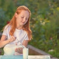 Fresh milk pours from a jug into a glass a red-haired girl. Warm sunlight illuminates the girl and the milk on the table. Glass of Royalty Free Stock Photo