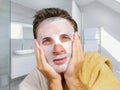 Young attractive and happy gay man in bathroom using beauty paper facial mask touching his face looking to the mirror smiling in Royalty Free Stock Photo