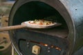 Fresh pizza on a wooden slide is pushed into a self-made recycling oven built from an old metal barrel, creative upcycling concept