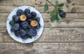 Fresh plums in bowl on wooden background with leaves and twig. Real photo from above. Halved plum with seed as decoration.