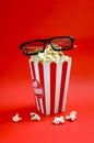 Fresh popcorn in a striped paper cup with 3d glasses on a red background.