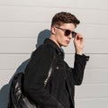 Fresh portrait fashionable young man in stylish denim black jacket in trendy sunglasses with hairstyle on sunny day near vintage Royalty Free Stock Photo