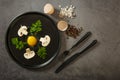 Fresh, raw chicken eggs with mushrooms and herbs, a fork and a knife on a black dish on a gray stone background, top view. Ingredi