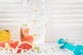 Fresh red grapefruits and grapefruit juice, dumbbell and measuring tape, on rustic white wooden table opposite the blinds.