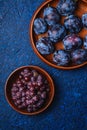 Fresh ripe plum fruits in brown wooden plate and grape berries in bowl on blue abstract background