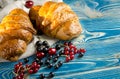 Fresh ruddy croissants with berries lie on a wooden table next to fresh black currant berries, red currants, cherries