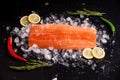 Fresh salmon fillet on a black background on chopped ice with sprigs of rosemary, slices of lemon, green and red chili peppers