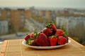 Fresh scarlet strawberries on a white plate stands on a bamboo napkin against the background of the blurry panorama of the city