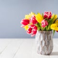 Fresh spring yellow and pink tulips bouquet in blue vase standing on white wooden table with gray background. Festive Royalty Free Stock Photo