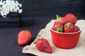 Fresh strawberries in mini red bowl on hessian jute. White and purple flowers in a decorative wooden crate. Black background