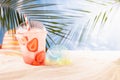 Fresh strawberry beverage with ice, straw in plastic glass on sandy tropical beach with sea view, palm leaves, beach toy.