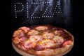 Fresh tasty Italian pizza on a black background with a spectacular light inscription `pizza` and smoke.