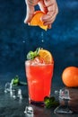 Fresh tropical cocktail with orange, mint and ice in glass on dark blue background. Man hand squeezes orange juice from tropical Royalty Free Stock Photo