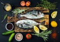 Fresh uncooked dorado or sea bream fish with lemon, herbs, oil, vegetables and spices on rustic wooden board over black