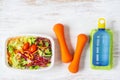 Fresh vegetable salad in lunch box with orange dumbbells excercise equipment and energy water drink on white rusty wood background Royalty Free Stock Photo