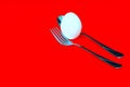Fresh white egg balanced between fork and a spoon on red background