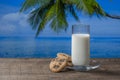 Fresh white milk in a glass with cookies on a wooden table with sea water, coconut palm tree and blue sky background on sunny Royalty Free Stock Photo
