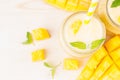 Freshly blended yellow mango fruit smoothie in glass jars with straw, mint leaves, mango slices, close up, top view. White wooden