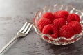 Freshly picked raspberries in a small glass bowl with a fruit fork to the side on a gray background