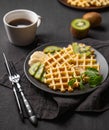 A freshly prepared waffles with kiwi and banana slices on a gray plate with a cup of coffee and a napkin on a dark background