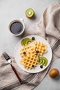 A freshly prepared waffles with kiwi, blueberries and banana slices on a white plate with a cup of coffee and a napkin on a light