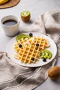 A freshly prepared waffles with kiwi, blueberries and banana slices on a white plate with a cup of coffee and a napkin on a light