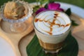 Glass of hot caramel macchiato coffee with cookie and orchid flower on wooden plate in coffee shop of Thailand.