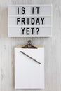 `Is it friday yet?` words on a modern board, clipboard with blank sheet of paper on a white wooden background, top view. Overhea
