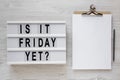 `Is it friday yet?` words on a modern board, clipboard with blank sheet of paper on a white wooden background, top view. Overhea