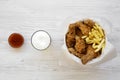 Fried chicken drumsticks, spicy wings, French fries and chicken fingers in paper box, sauce and cold beer over white wooden table,
