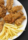 Fried chicken drumsticks, spicy wings, French fries, chicken fingers on white plate over black background, top view. Flat lay, Royalty Free Stock Photo