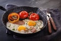 Fried eggs with bacon and tomatoes on an old cast iron pan and cutlery on a gray table. Royalty Free Stock Photo