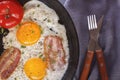 Fried eggs with bacon and tomatoes on an old cast iron pan and cutlery on a gray table. Royalty Free Stock Photo